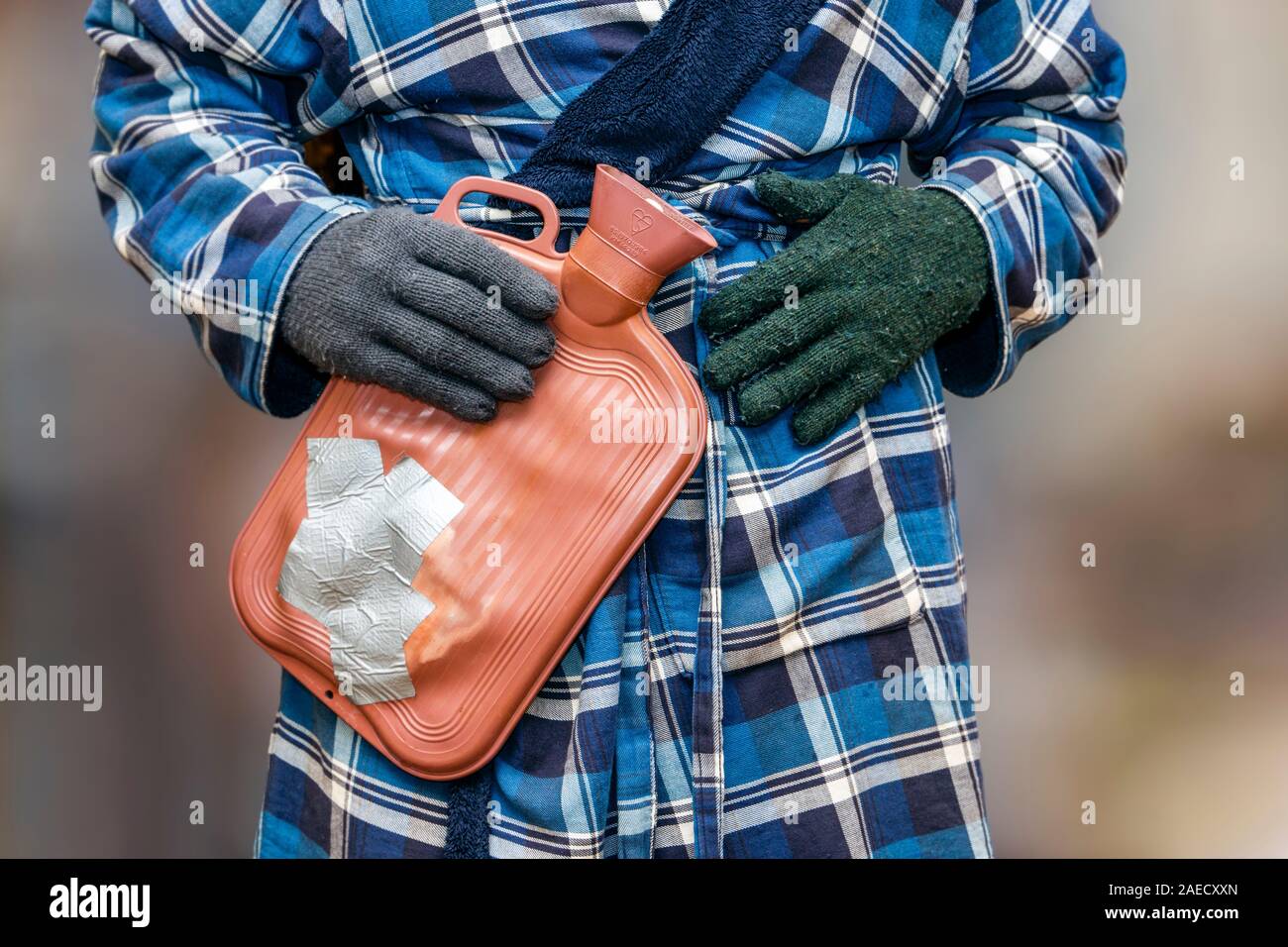 Ein verletzlicher Rentner in Morgenmantel und ungetrübten Wollhandschuhen, der eine geflickte Wärmflasche hält und versucht, sich warm zu halten, während der Treibstoff in die Höhe schnellen lässt Stockfoto