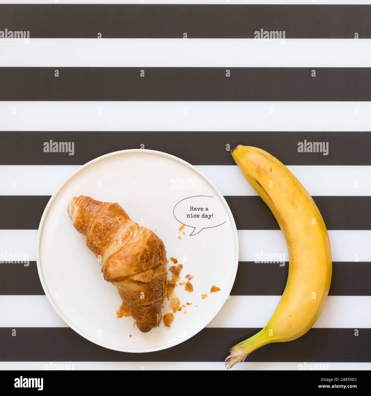 Bio Banane auf schwarze und weiße Streifen Hintergrund. Ansicht von oben. Französische Croissant auf einem weißen Teller mit Vorzeichen haben Sie einen schönen Tag. Leckeres Frühstück. Stockfoto