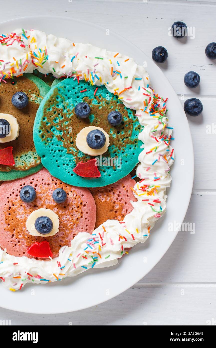 Mehrfarbige Pfannkuchen mit Früchten und Schlagsahne für Kinder auf eine weiße Platte, weisser Hintergrund. Stockfoto