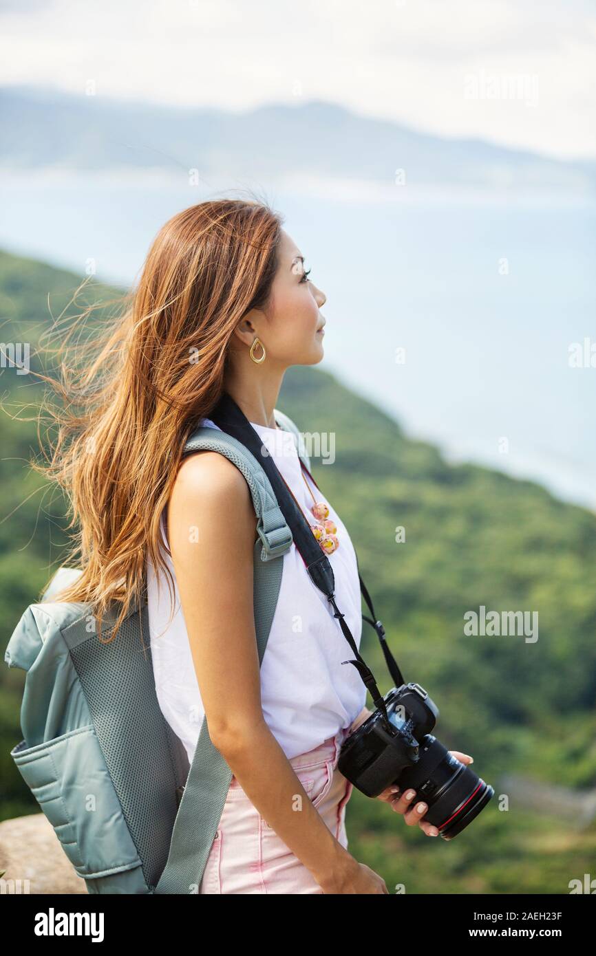 Japanische Frau, die Rucksack und Halten der Kamera steht auf einer Klippe, auf das Meer im Hintergrund. Stockfoto