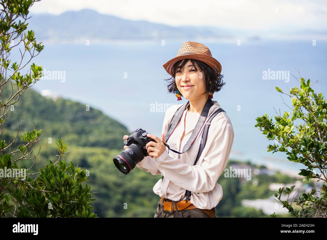 Japanische Frau mit Hut und mit Rucksack und Kamera steht auf einer Klippe, auf das Meer im Hintergrund. Stockfoto