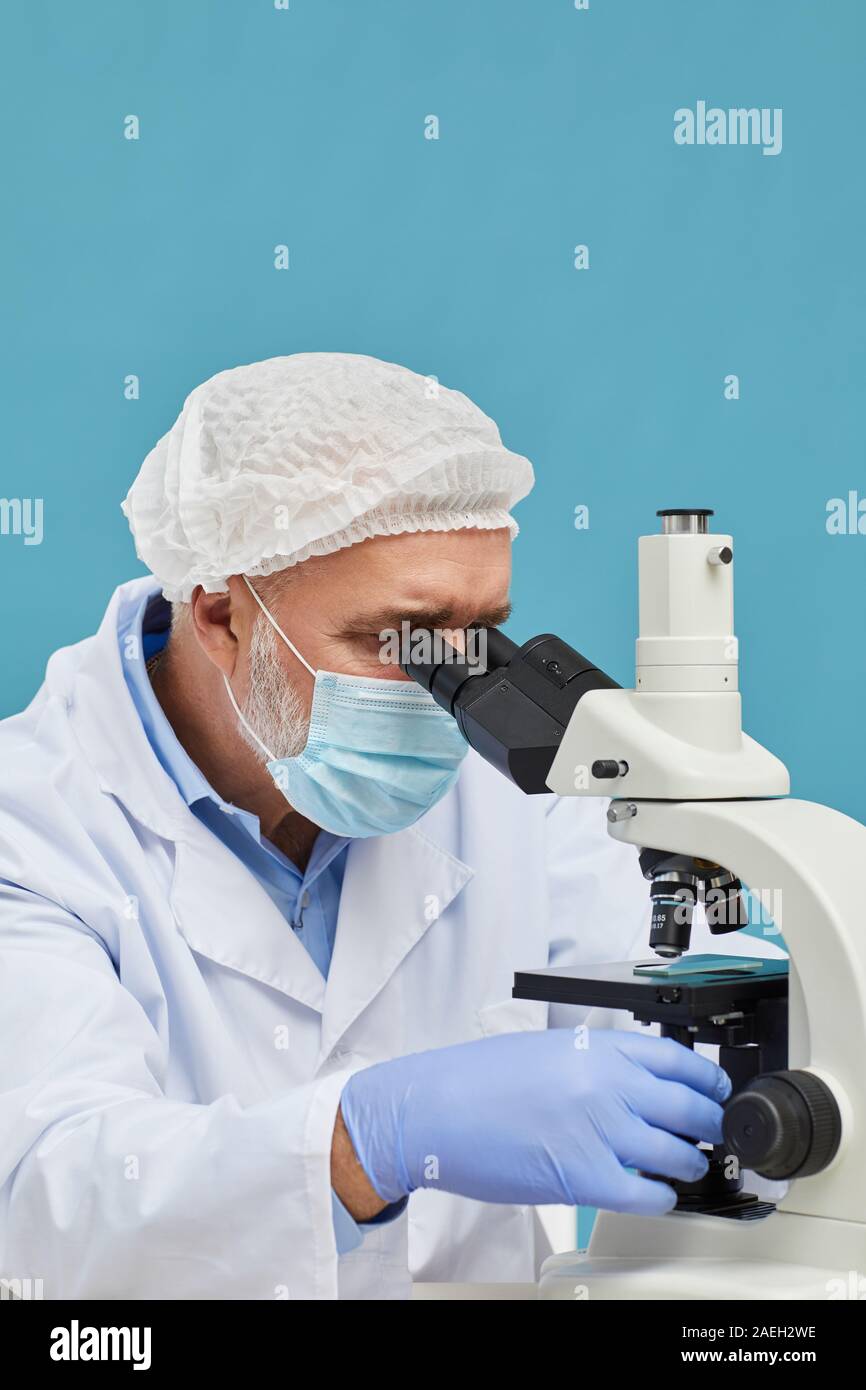Reifen Arzt durch das Tragen von schützender maske und handschuhe Prüfung der Probe durch das Mikroskop auf blauem Hintergrund isoliert Stockfoto