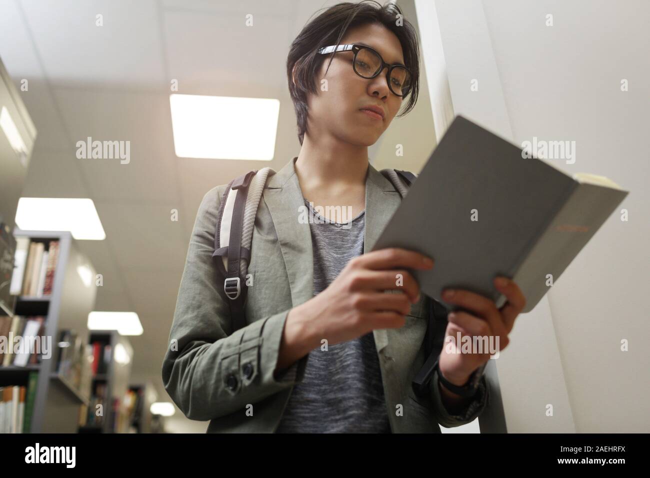 Asiatische junge Mann in Brillen und mit Rucksack auf dem Rücken, ein Buch zu lesen, während man in der Bibliothek Stockfoto