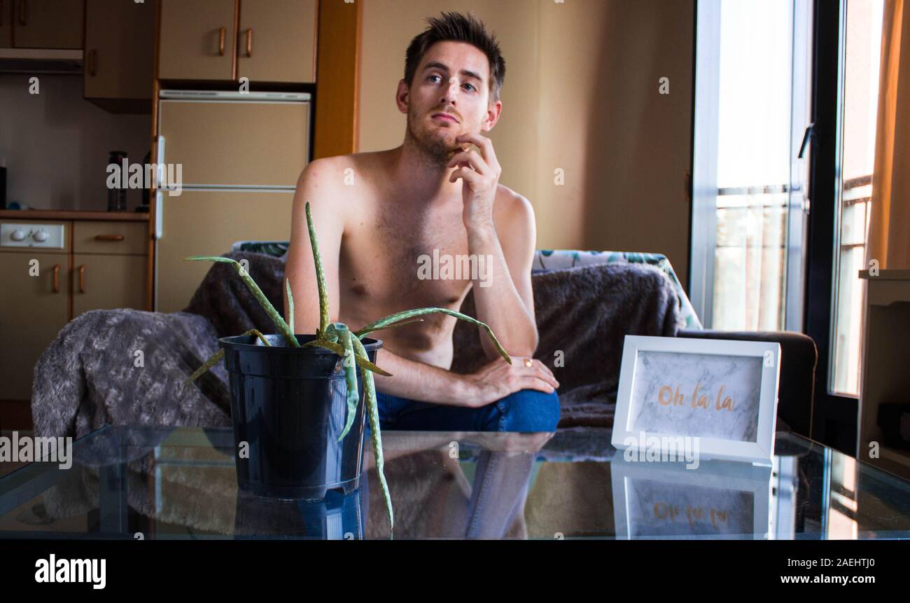 Ein intelligenter junger Mann sitzt auf einem Sofa ohne ein Hemd am Fenster suchen Stockfoto