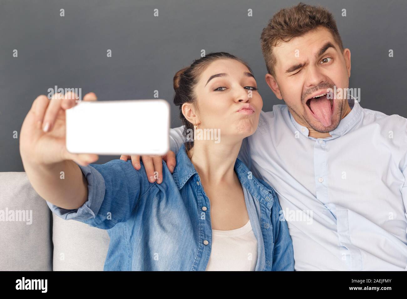 Relationnship Konzept. Junges Paar umarmen Sitzen auf einem Sofa studio isoliert auf Grau unter selfie auf dem Smartphone Grimassen zu Kamera verspielt Stockfoto