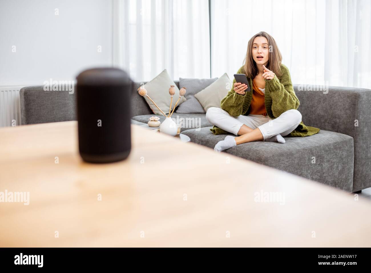 Junge Frau controlling home Geräte mit Sprachbefehlen, Gespräch mit einem smart Spalte, während auf der Couch zu Hause entspannen Stockfoto