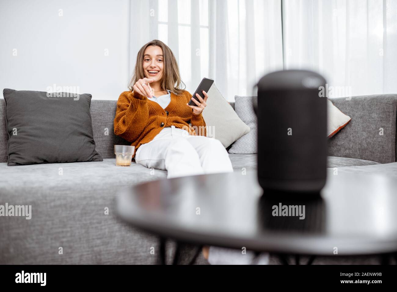 Junge fröhliche Frau controlling home Geräte mit Sprachbefehlen, Gespräch mit einem smart Spalte, während auf der Couch zu Hause entspannen Stockfoto
