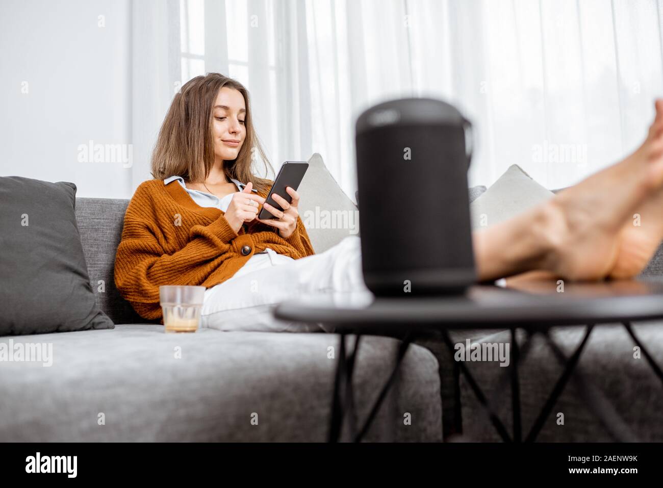 Junge fröhliche Frau controlling home Geräte mit Sprachbefehlen, Gespräch mit einem smart Spalte, während auf der Couch zu Hause entspannen Stockfoto