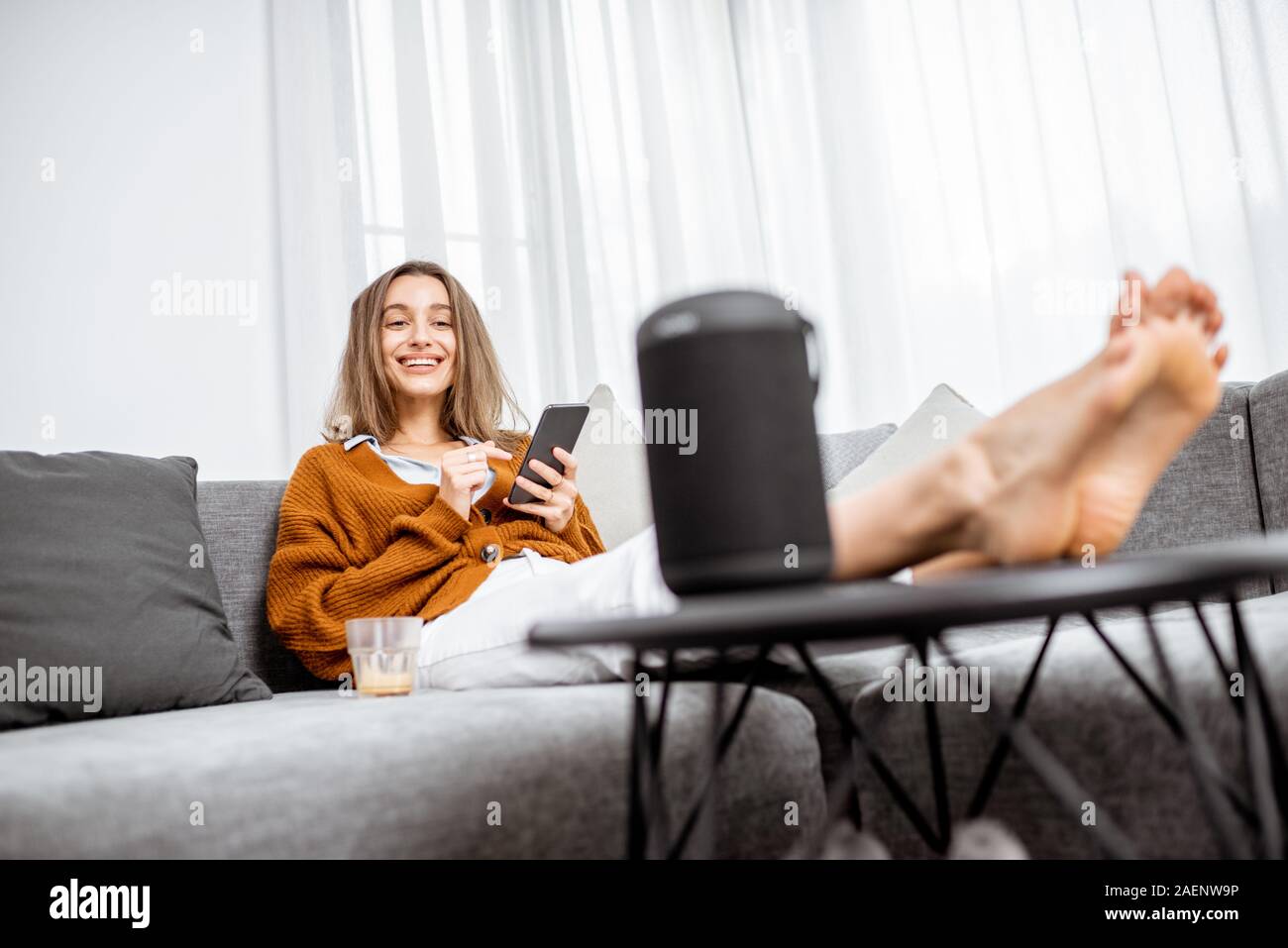 Junge fröhliche Frau controlling home Geräte mit Sprachbefehlen, Gespräch mit einem smart Spalte, während auf der Couch zu Hause entspannen Stockfoto