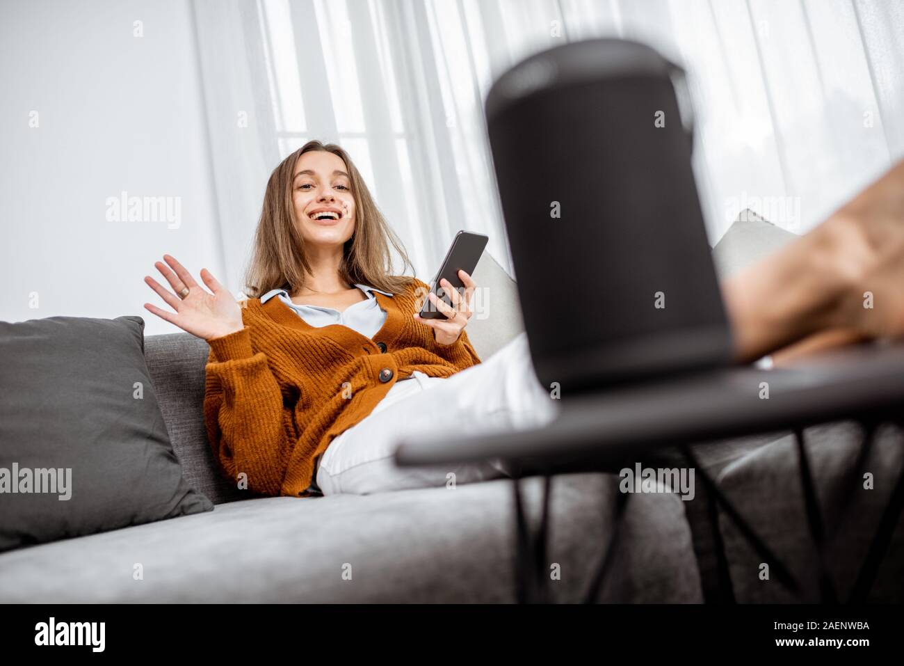 Junge fröhliche Frau controlling home Geräte mit Sprachbefehlen, Gespräch mit einem smart Spalte, während auf der Couch zu Hause entspannen Stockfoto