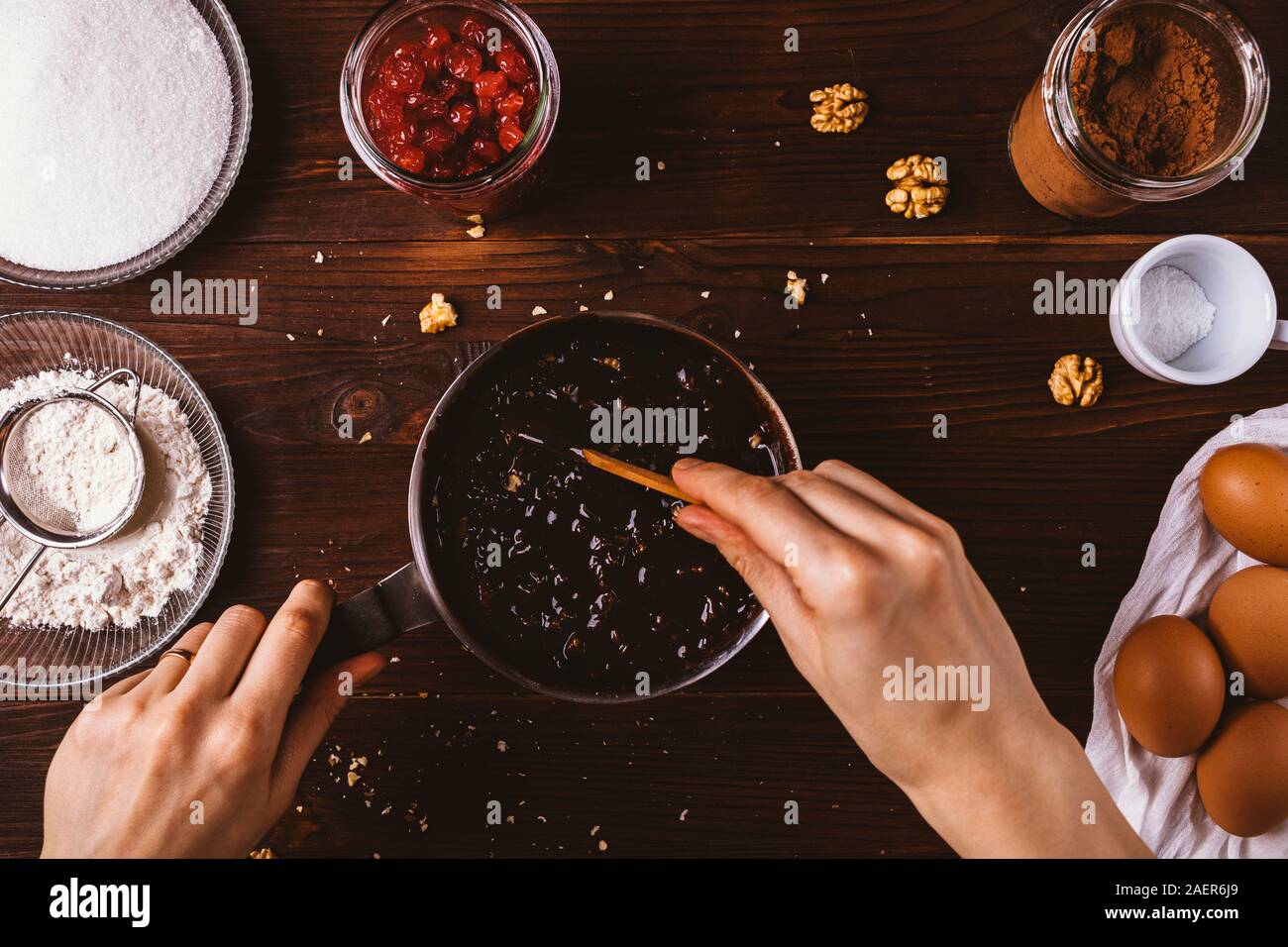 Die Hand des Weiblichen Mischung geschmolzener Schokolade mit gehackten Walnüssen in Kochtopf unter anderen Zutaten auf dunklen Holztisch. Draufsicht der Teig für Stockfoto