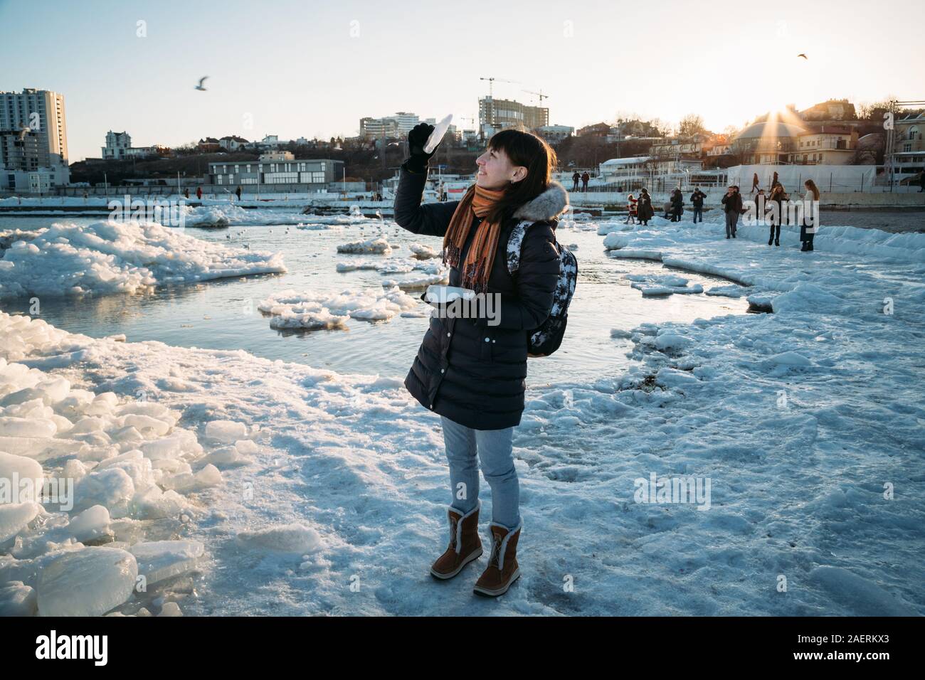 Odessa, Ukraine - Februar, 16 2017: junge lächelnde Frau, die auf gefrorenen Küste des Schwarzen Meeres mit Eis holding Stück Eis bei Sonnenuntergang fällt. Stockfoto