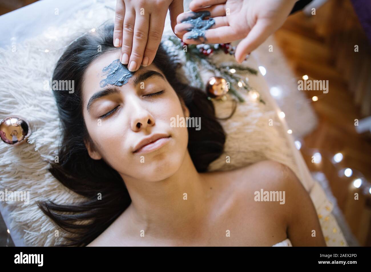 Therapeuten, die Maske auf Girl's Stirn im SPA Salon. Weibliche Hände Anwendung Holzkohle Maske auf das Gesicht einer Frau liegend auf Bett mit Lichter und weihnachtliche Stockfoto