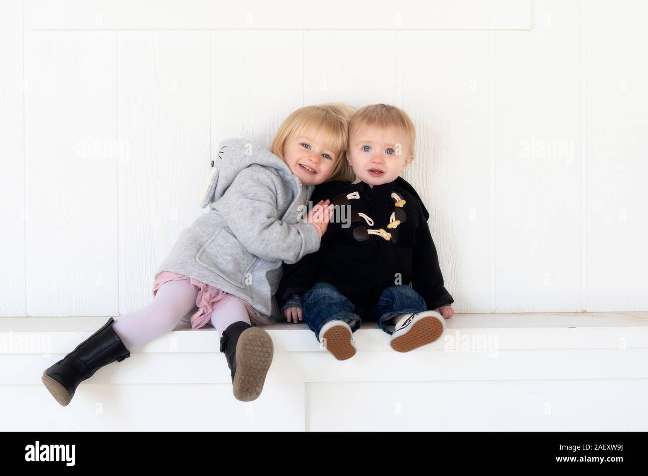 Blonde, blauäugige, Kleinkind Junge und Mädchen Geschwister zusammen sitzen auf einer Bank in warme Winterkleidung mit einem natürlichen weißen Holz Hintergrund gekleidet Stockfoto