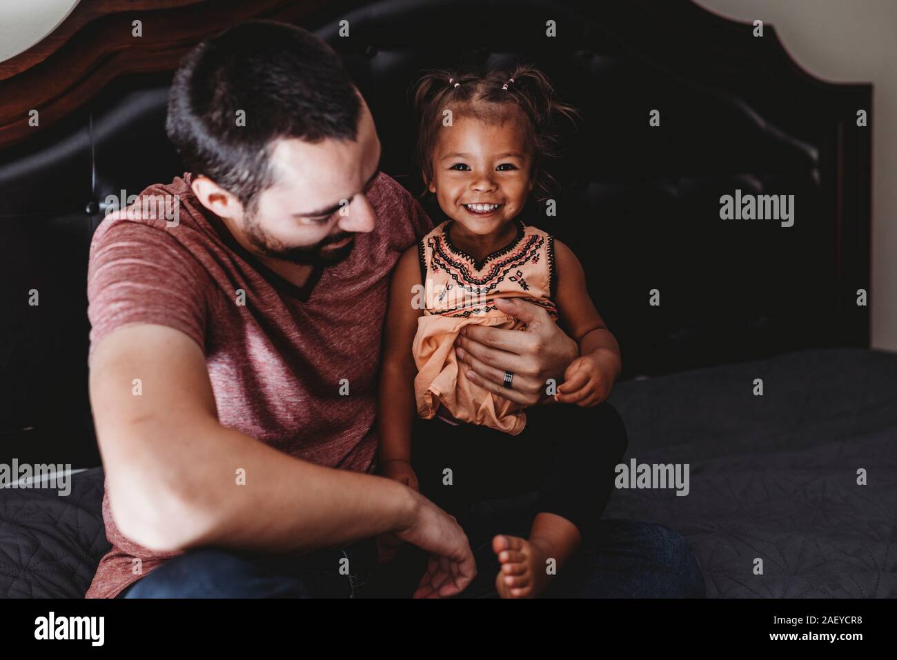 Schöne 2-jährigen gemischtrassigen Mädchen sitzen auf dem Bett mit Dad lächelnd Stockfoto