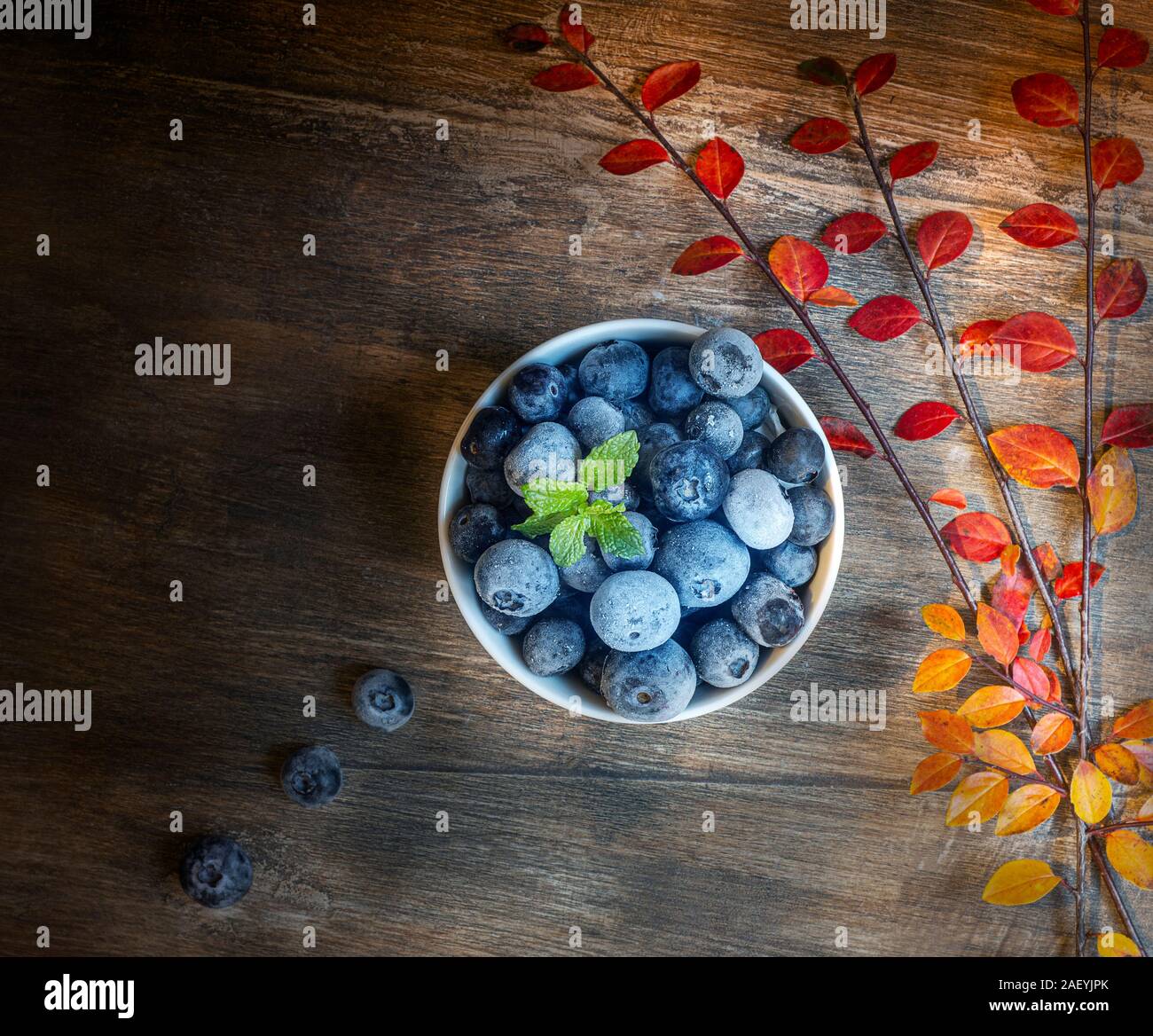 Gefrorene Heidelbeeren in einer weissen Schüssel mit Blätter im Herbst wie kostenlose Farben auf einem strukturierten hölzernen Tisch. Stockfoto