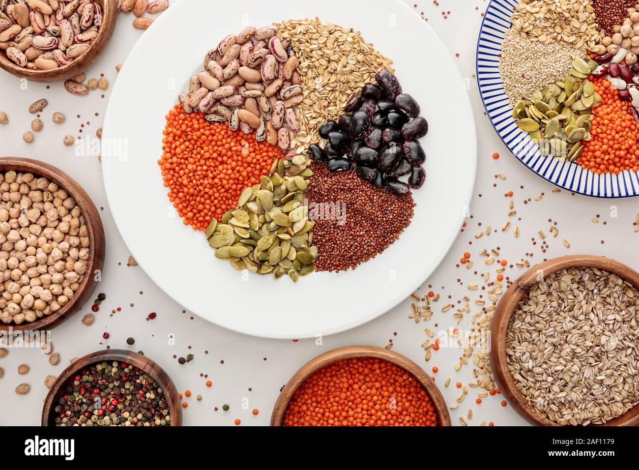 Blick von oben auf die Teller und Schalen mit roh Linsen, Quinoa, Haferflocken, Bohnen, Pfefferkörner und Kürbiskerne auf Marmor Oberfläche mit verstreuten Körner Stockfoto