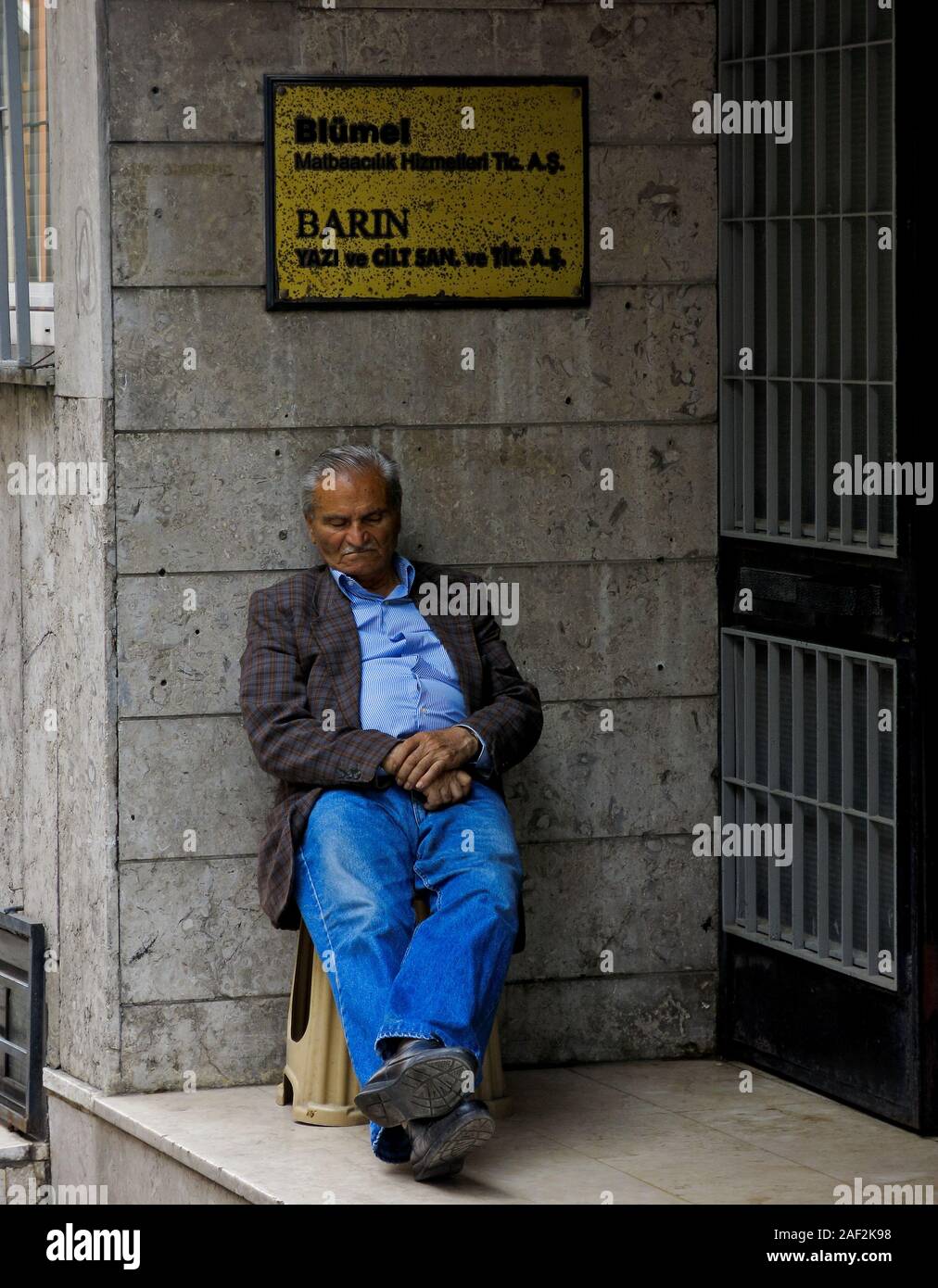 Ein älterer Mann lehnt sich an eine Wand und snoozes entlang der Straße Stockfoto