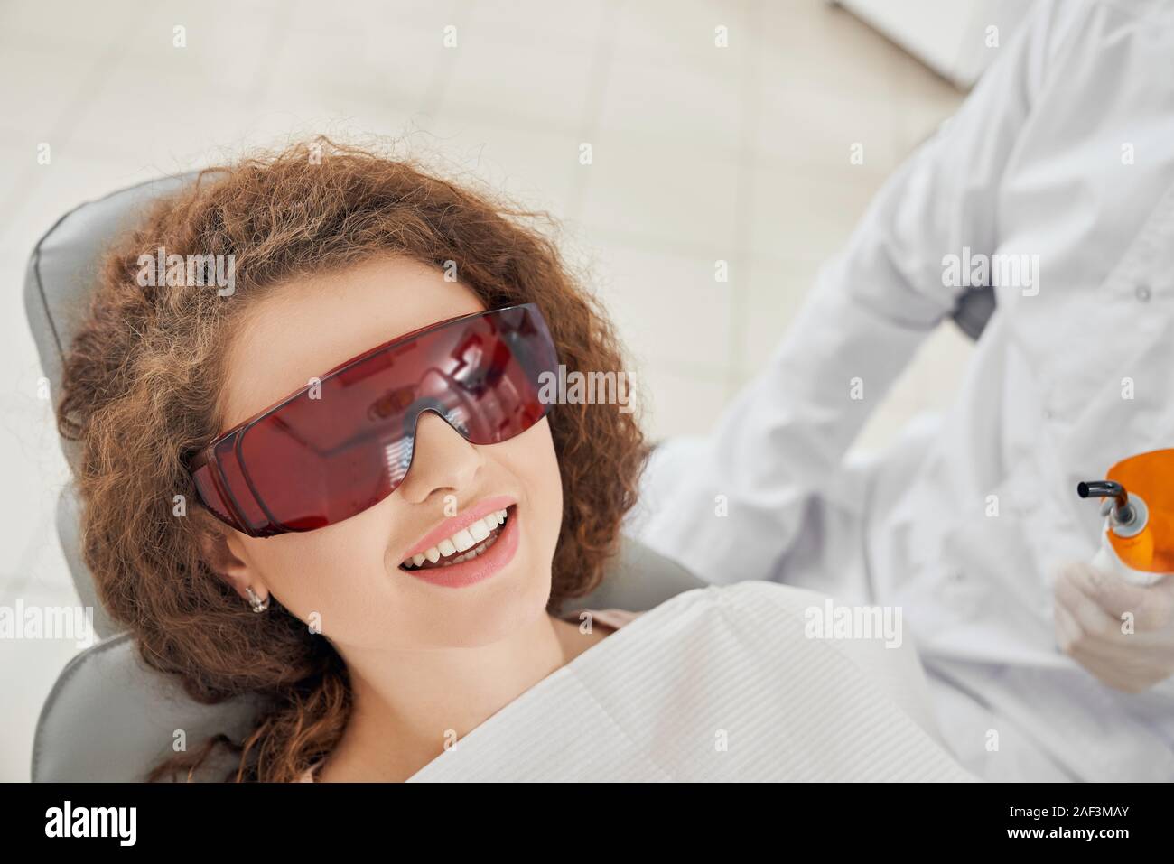Blick von oben der schönen Frau liegend auf zahnmedizinischen Stuhl in eine Schutzbrille. Curly Patientin lächelnd, während sie die Verfahren. Konzept der Zahnmedizin und die Zähne. Stockfoto