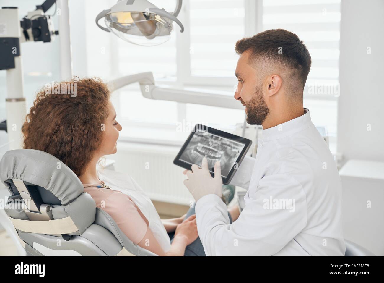 Ansicht von der Seite des männlichen Zahnarzt arbeiten in der modernen zahnmedizinischen Klinik. Professionelle Mann im weißen Uniform halten Tablet mit x-ray Bild von Zähnen und Übersicht zu den weiblichen Patienten. Konzept der Zahnheilkunde. Stockfoto