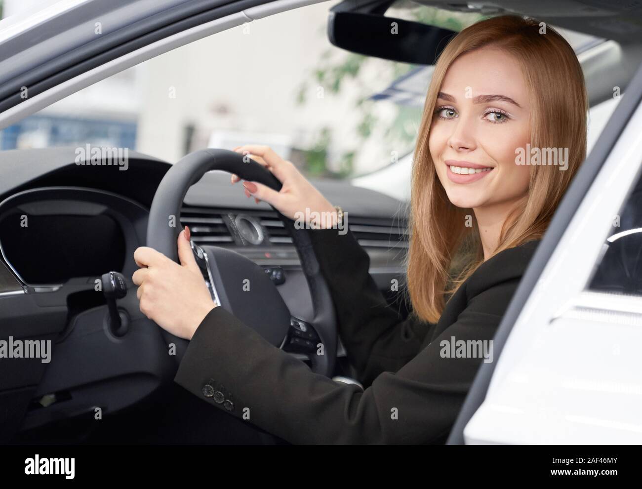 Blick von der offenen Tür von attraktiven weiblichen Kunden der Auto Salon in Weiß Auto sitzen. Lächelnde Blondine Testen neuer Auto, halten die Hände auf Rad und Kamera. Konzept der Auto kaufen. Stockfoto