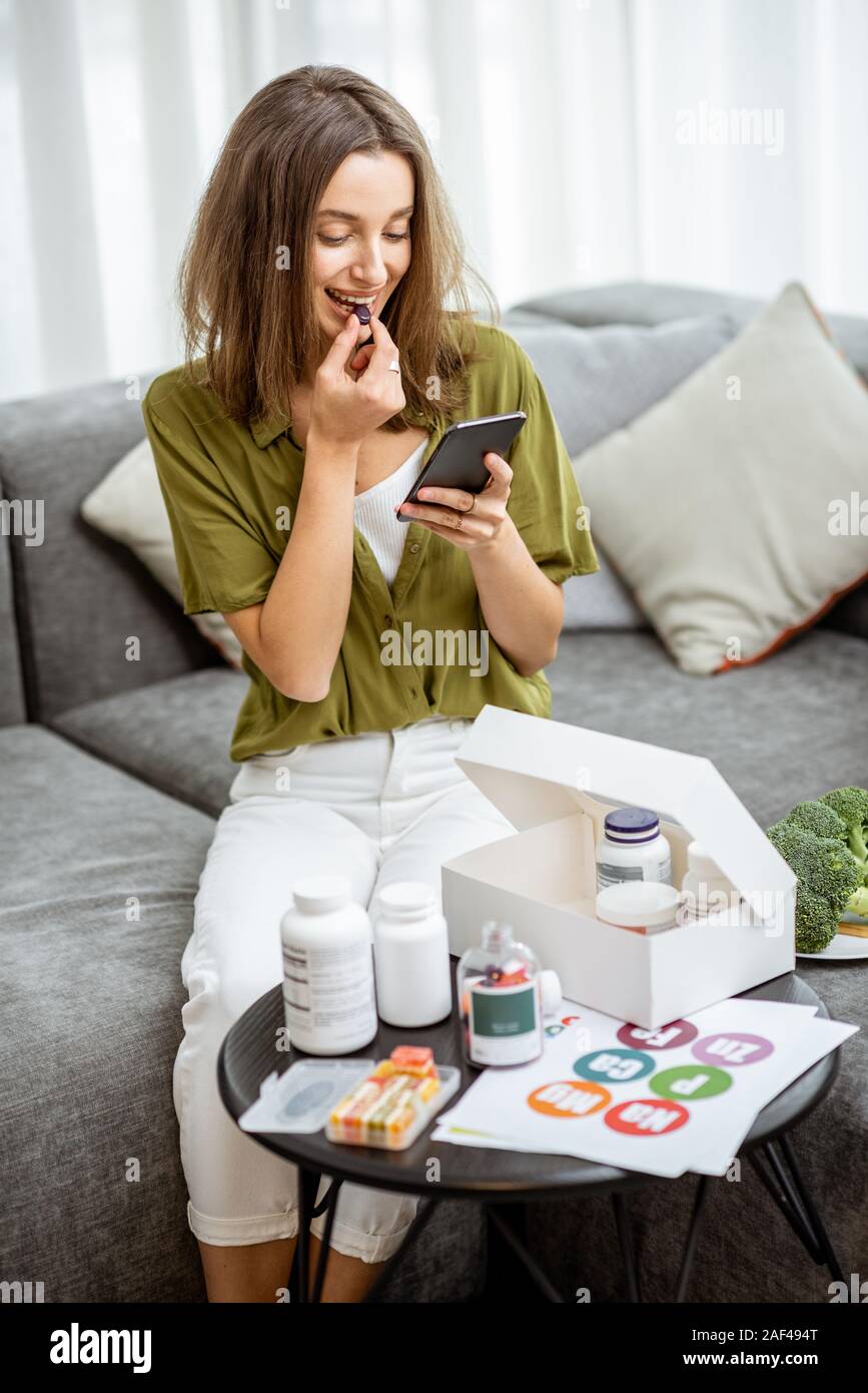 Frau Einnahme von Nahrungsergänzung in Form von Pillen, während mit Smart Phone, zu Hause zu sitzen. Konzept der individuellen online Auswahl von Nahrungsergänzungsmitteln. Präventive Medizin Stockfoto