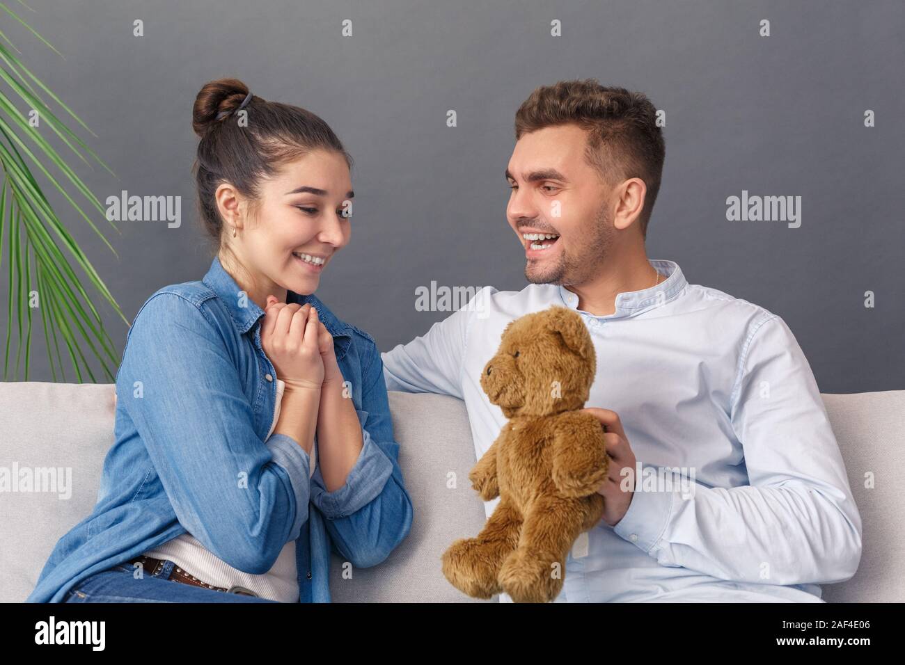 Relationnship Konzept. Junges Paar Mann, Teddybären zu lächelnde Frau glücklich sitzen auf einem Sofa studio isoliert auf Grau Stockfoto