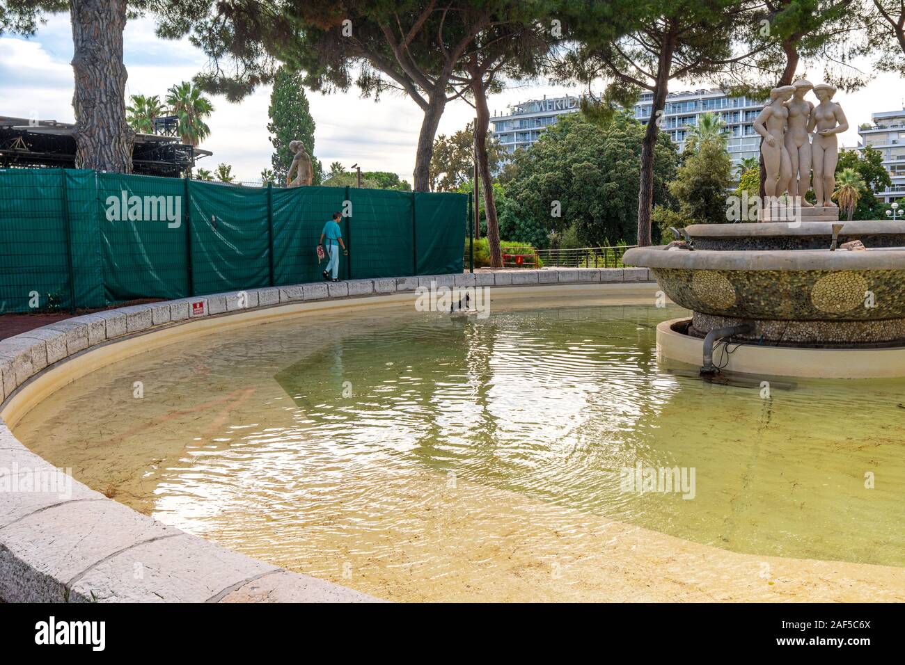 Eine Frau beobachtet, wie ihr kleiner Hund spielt im Wasser an die Drei Grazien Brunnen im Albert Park an der Französischen Riviera. Stockfoto