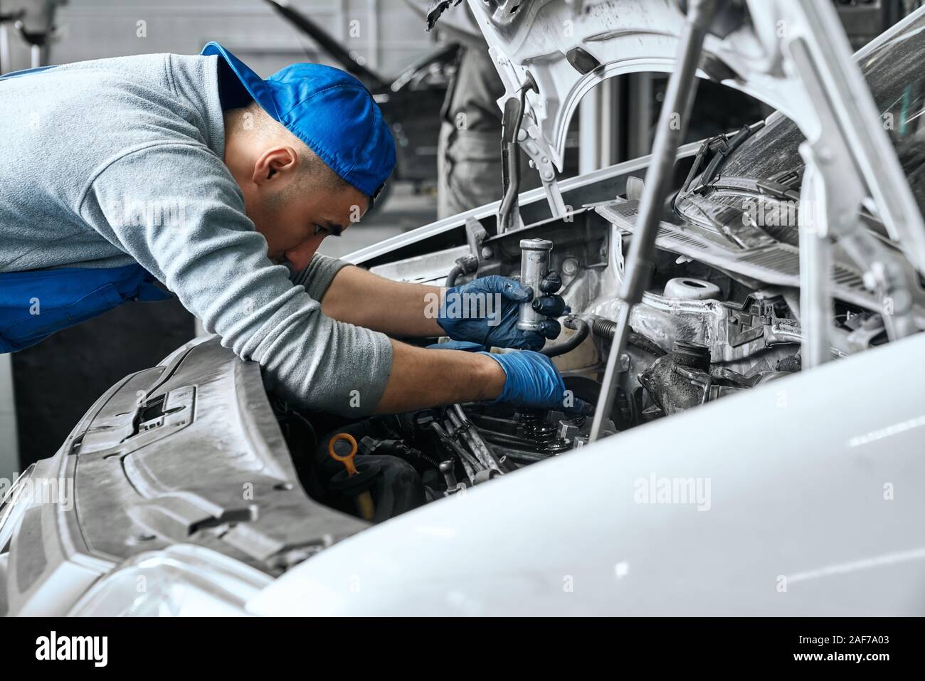 Männliche Auto fahrer tragen blauen Overall, Mütze und Handschuhe arbeiten unter der Haube von weißes Auto und aufmerksam prüfen Wartung des Motors bei Reparatur Stockfoto