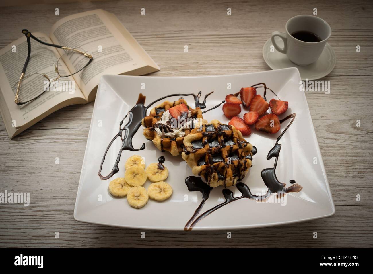 Leckere Waffeln mit Erdbeeren, Bananen und Schokolade mit einer Tasse Kaffee und einem Buch auf dem Tisch Stockfoto