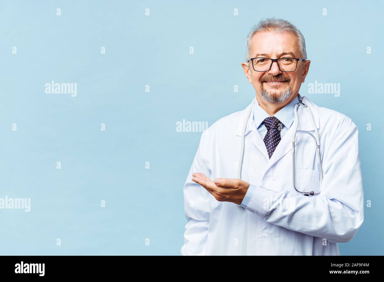Fröhliche reife Arzt posing und lächelnd an Kamera, Gesundheitswesen und Medizin. Auf blauem Hintergrund isolieren. Hand bei copy Raum zeigt. Stockfoto