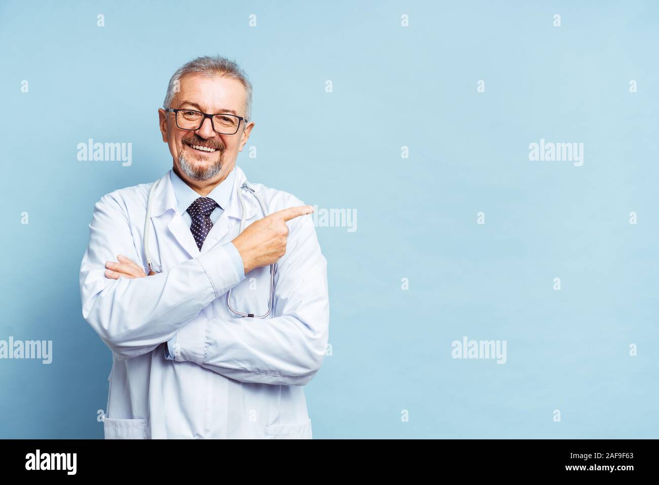 Fröhliche reife Arzt posing und lächelnd an Kamera, Gesundheitswesen und Medizin. Auf blauem Hintergrund isolieren. Hand bei copy Raum zeigt. Stockfoto