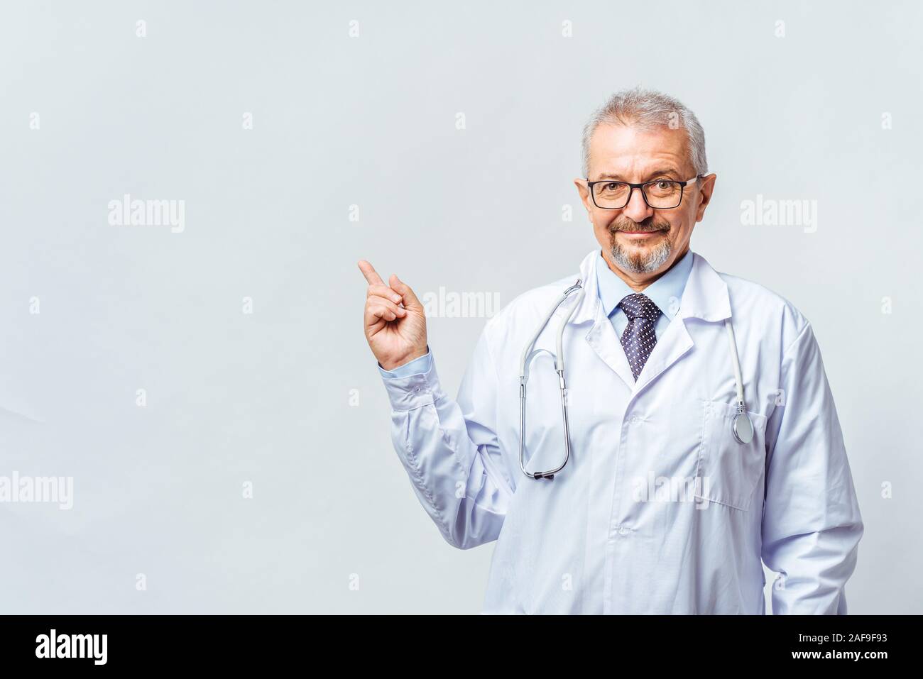 Fröhliche reife Arzt posing und lächelnd an Kamera, Gesundheitswesen und Medizin. Auf blauem Hintergrund isolieren. Hand bei copy Raum zeigt. Stockfoto