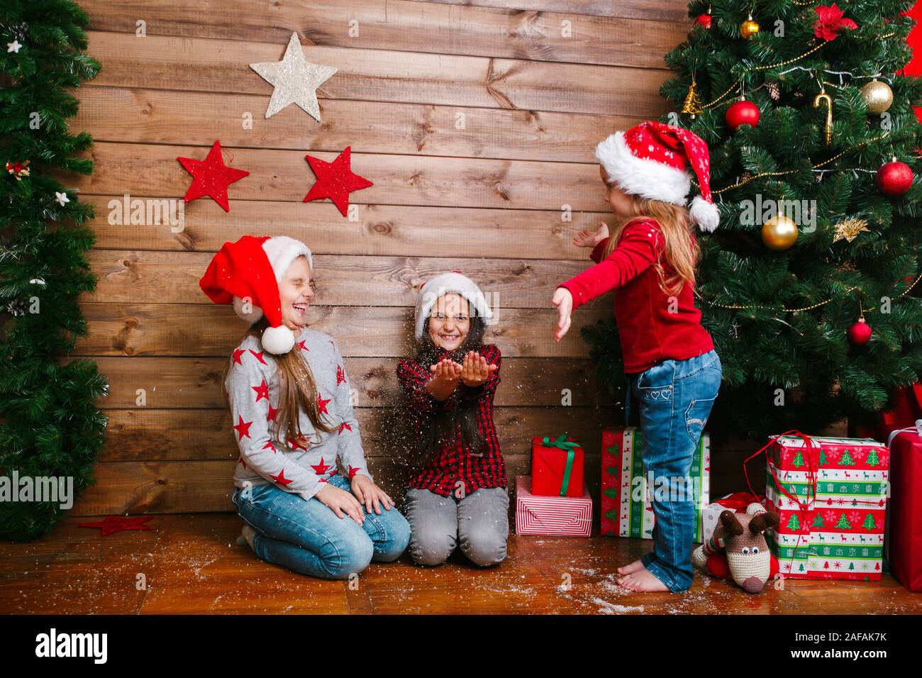 Drei glückliche Lustige kleine Schwestern in Santa Hüte mit Geschenken Spaß in der Nähe von Weihnachten Baum Stockfoto