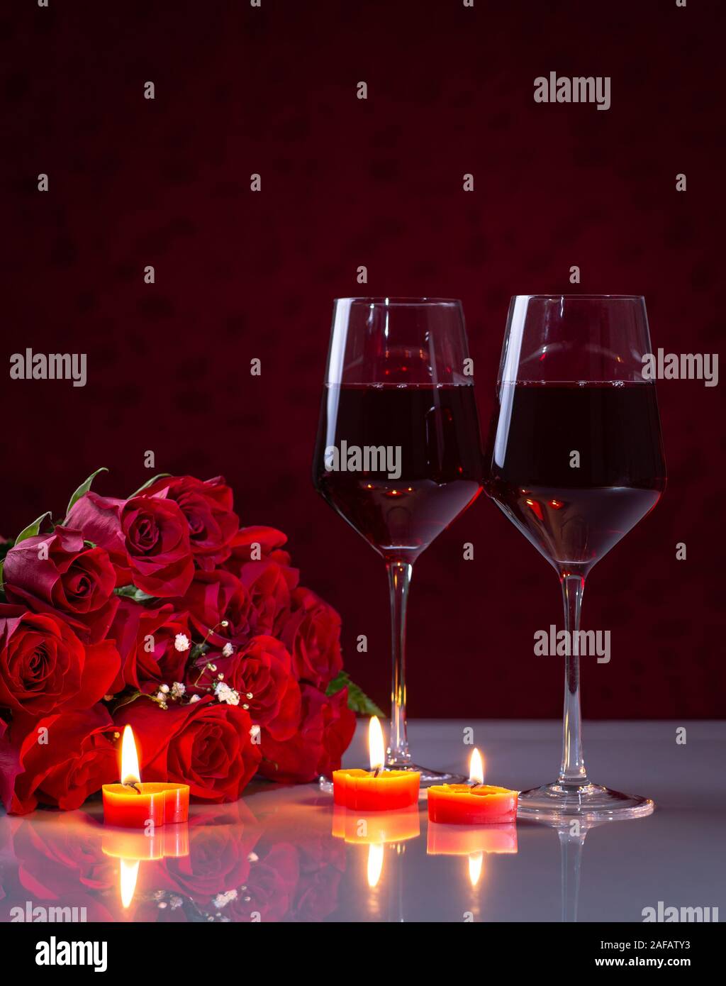 Blumenstrauß aus roten Rosen und zwei Gläser Wein mit brennenden Kerzen auf eine reflektierende weiße Tischplatte Stockfoto