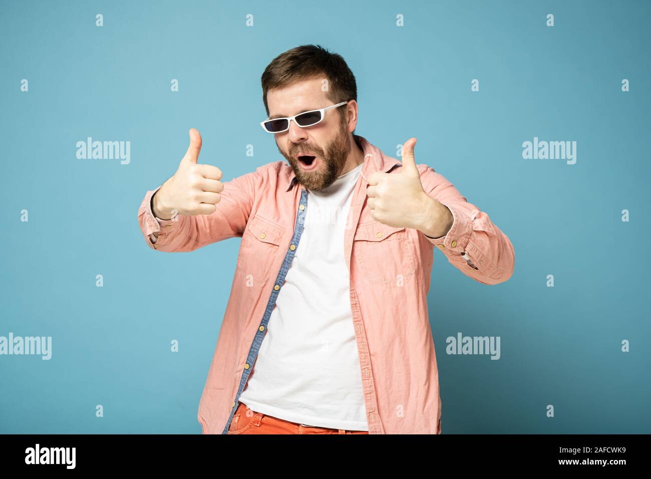 Seltsam, bärtigen Mann in Sonnenbrille Gesten zeigen Daumen bis auf zwei Hände, öffnet sich Mund und macht einen seltsamen Gesichtsausdruck. Stockfoto