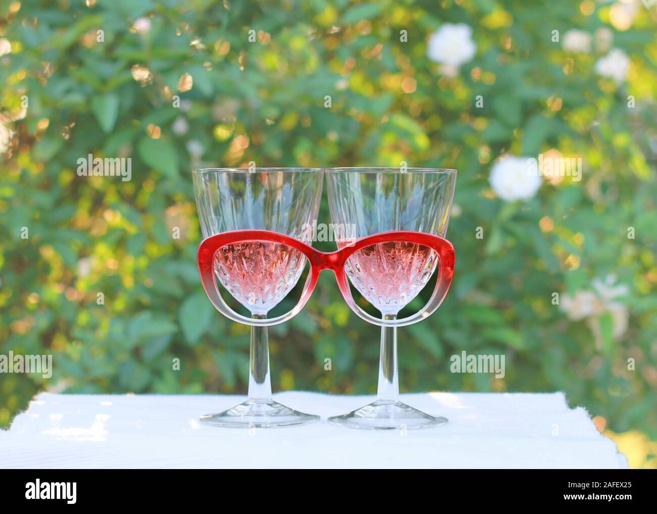 Eine skurrile Einstellung von zwei Weingläser mit Rose Wein und rote Brillen steht vor einem Rosenbusch. Stockfoto