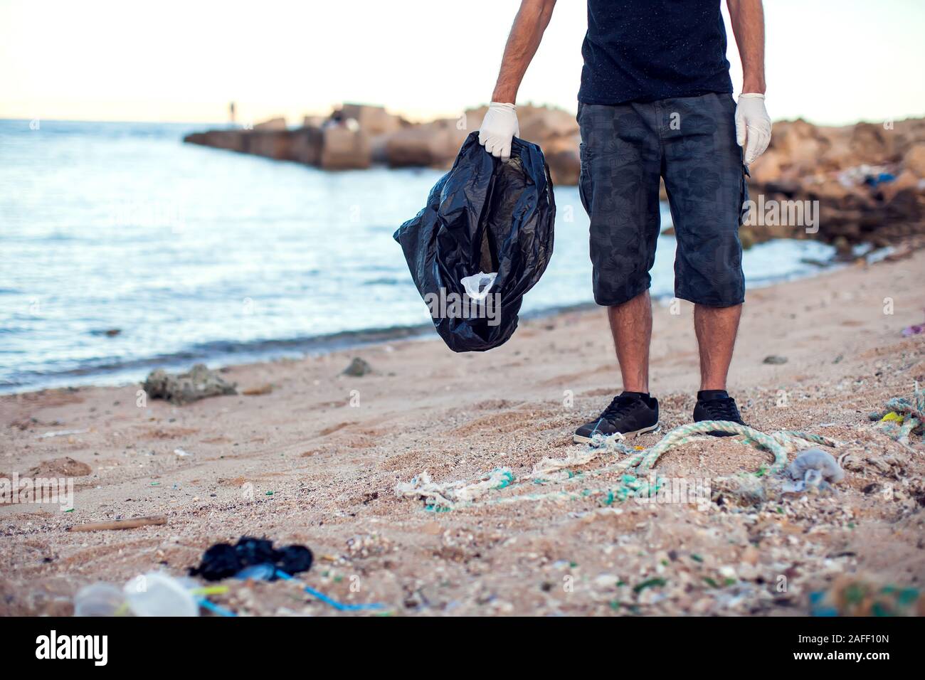 Mann in dunklem Shirt und Shorts mit weißen Handschuhen und großen schwarzen Paket Garbage Collecting am Strand. Umweltschutz und Planeten Verschmutzung co Stockfoto