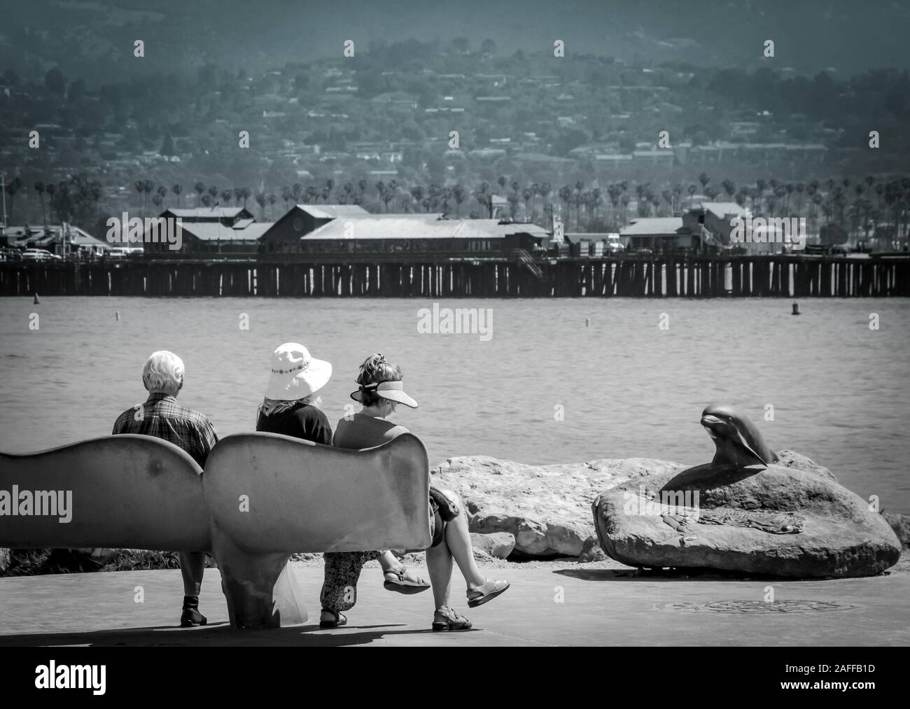 Die Leute sitzen auf der Whale Tale Bänke mit Blick auf den Hafen von Santa Barbara mit einer Bronze dolphin Skulptur und Sterns Wharf Santa Barbara, CA, USA. Stockfoto