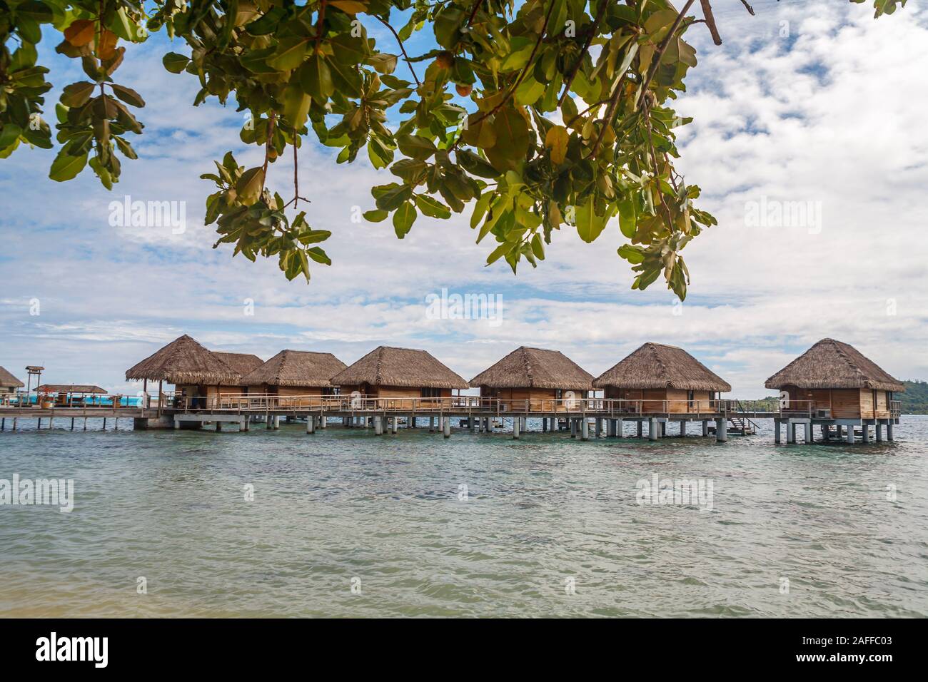 Blick auf eine Reihe von typischen strohgedeckten Beachside Überwasser Bungalows durch einen von Palmen gesäumten Strand auf Bora Bora, einer kleinen Insel im Südpazifik in Französisch-Polynesien Stockfoto