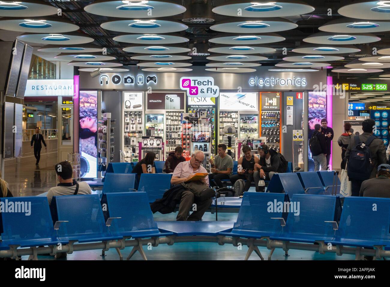 Passagiere sitzen auf Sitzen vor einem Tech & Technik Shop im Terminal 4 S von Adolfo Suárez Flughafen Barajas, Madrid, Spanien Fliegen Stockfoto