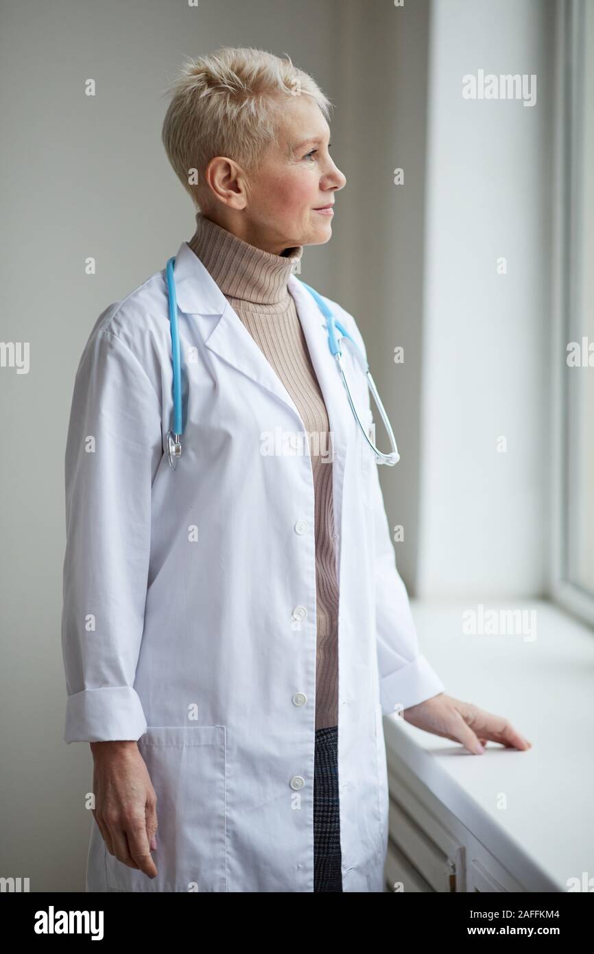 Taille bis Portrait von reife weibliche Arzt stehen durch Fenster in Klinik und weg schauen nachdenklich Stockfoto