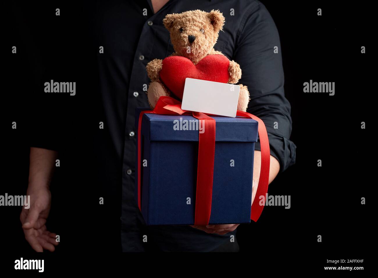 Erwachsener Mann in einem schwarzen T-Shirt hält die blaue Kästchen mit einem roten Band gebunden und befindet sich auf der Spitze eines braunen Teddybär mit Herz, Konzept der congratulatio Stockfoto