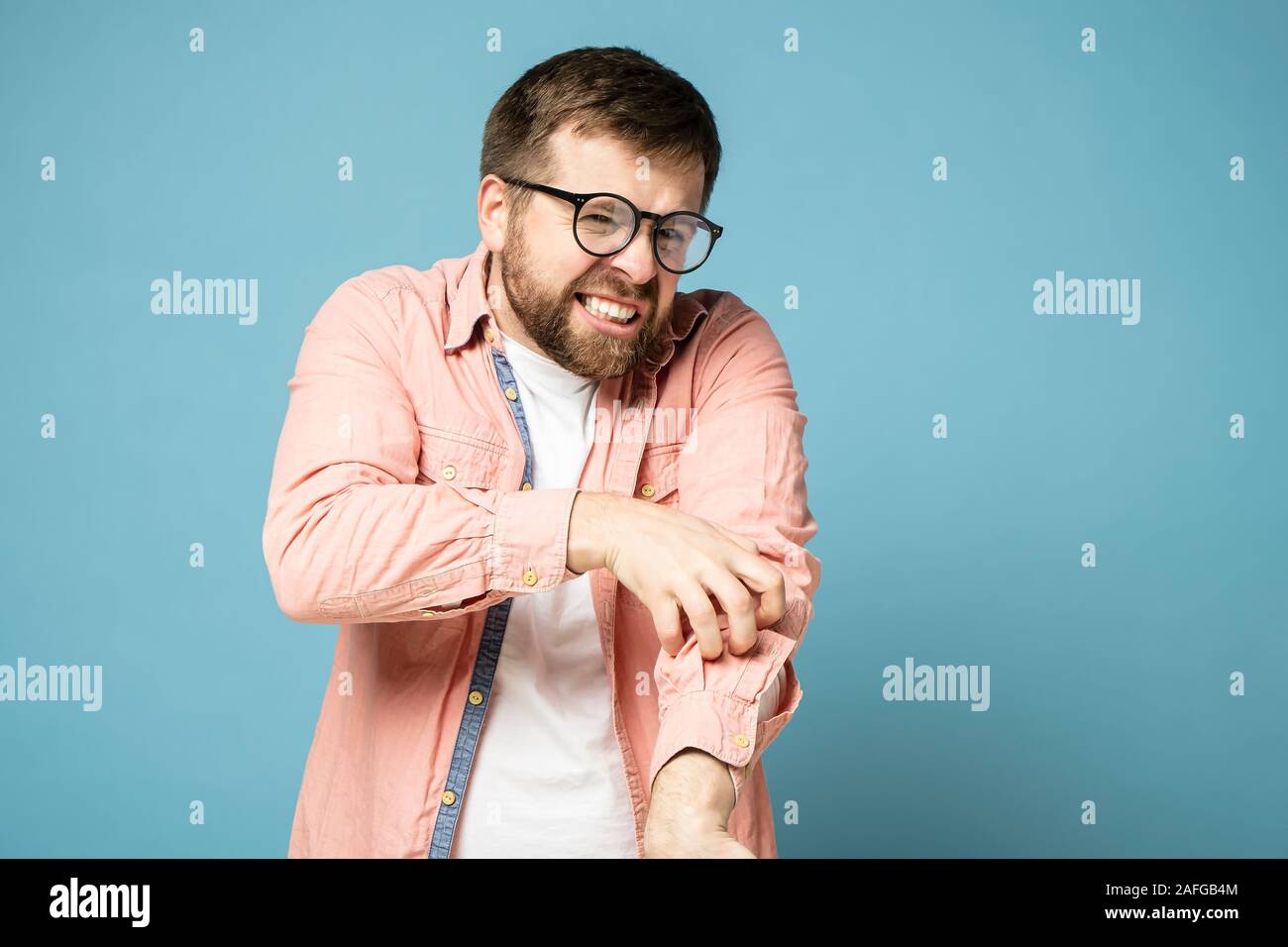 Zeitform bärtiger Mann mit Brille Kratzer juckende Haut auf der Hand durch ein Hemd, schmerzlich grinst Zähne und sieht in die Kamera. Stockfoto