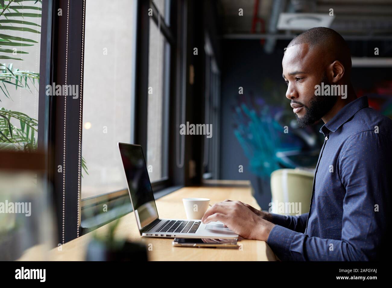 Mitte der erwachsenen männlichen Schwarzen kreative Sitzen durch Fenster in einem Büro, Sozialraum mit einem Laptop, Seitenansicht Stockfoto