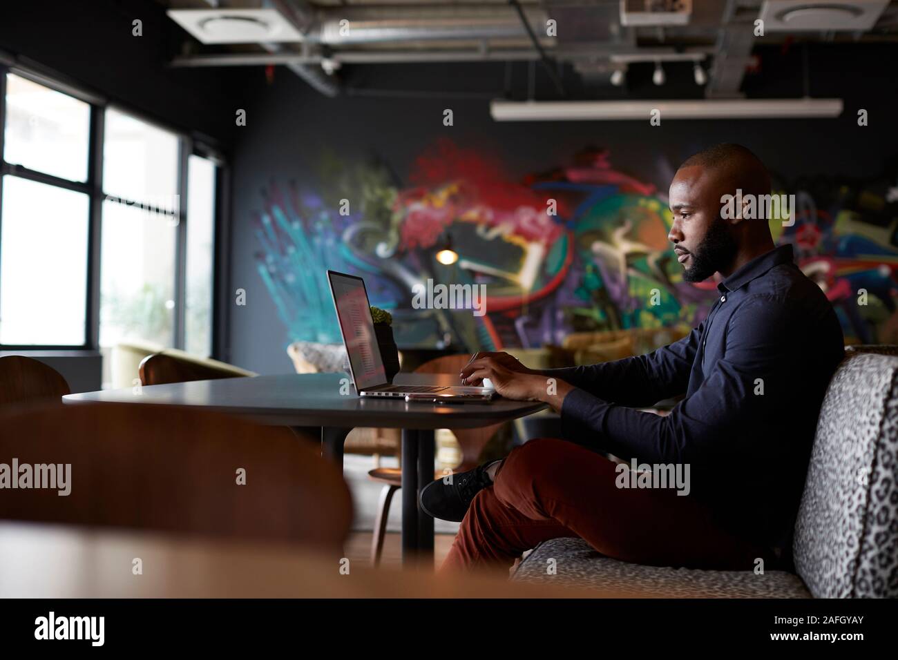 Mitte der erwachsenen männlichen Schwarzen kreative Sitzen am Tisch im Büro Essbereich mit einem Laptop, Seitenansicht Stockfoto