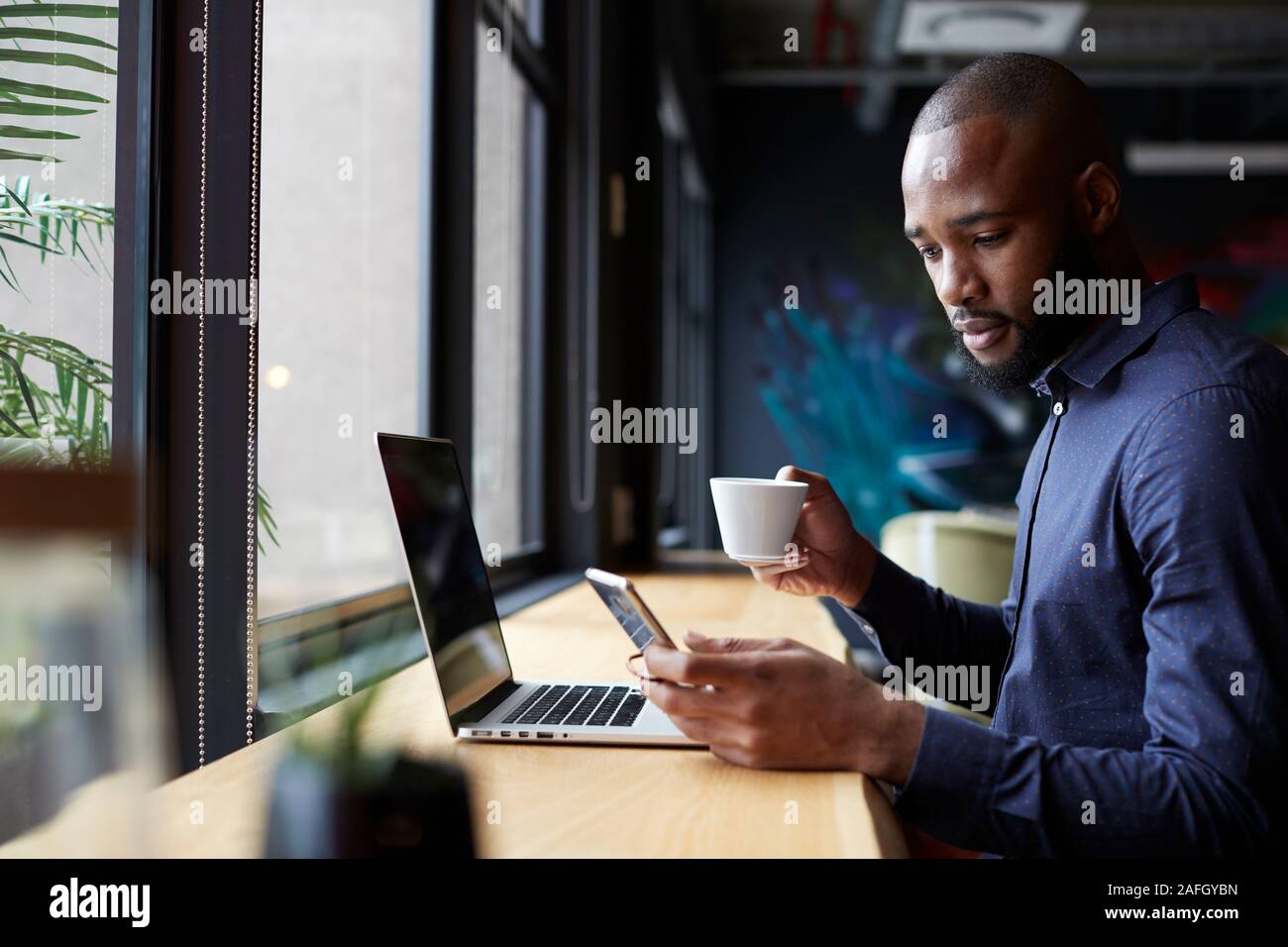 Mitte der erwachsenen männlichen Schwarzen kreative sitzt am Fenster Kaffee, mit einem Laptop und Smartphone, Seitenansicht Stockfoto