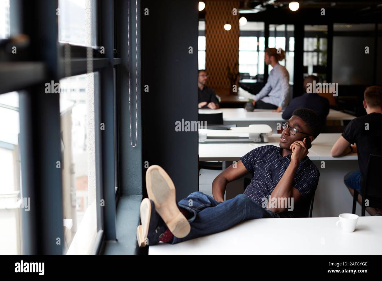 Junge schwarze Männer kreative sitzt in einem Büro mit seinen Füßen auf dem Schreibtisch mit Telefon, bis in der Nähe Stockfoto