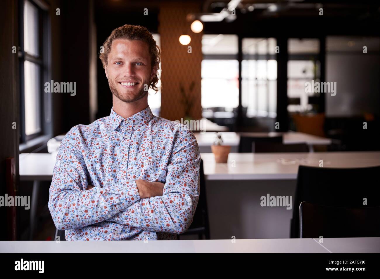 Tausendjährige weißen männlichen Kreative sitzt in einem Büro mit gekreuzten Armen lächelnd in die Kamera, in der Nähe Stockfoto