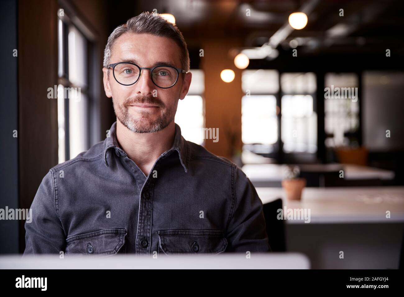 Im mittleren Alter weiße männliche Kreative sitzt in einem Büro lächelnd in die Kamera, Kopf und Schultern, in der Nähe Stockfoto